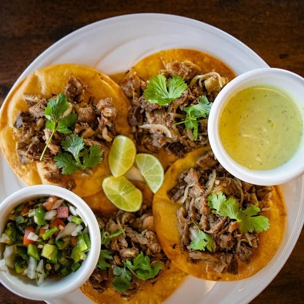 Four street tacos with meat, cilantro, lime, pico de gallo, and green sauce on a white plate.