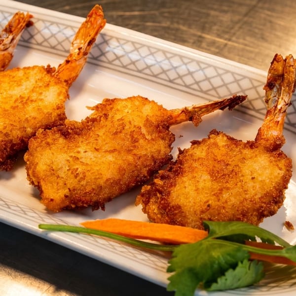 Four golden-brown fried shrimp with tails on, garnished with a carrot slice and cilantro.